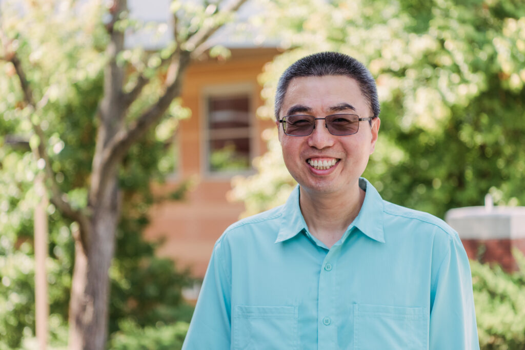 Portrait of man smiling outdoors wearing blue collared shirt and transition lens glasses darkened by the sun.