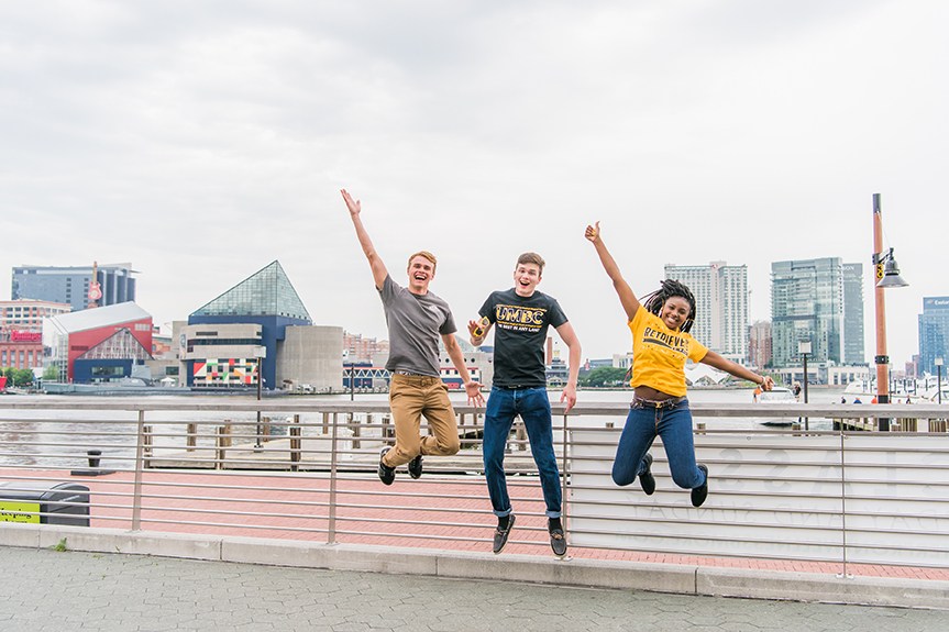 3 students wearing UMBC shirts jumping infront of baltimore habor