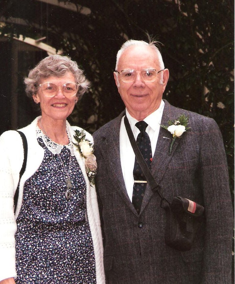 A man wearing a grey suit and glasses stands next to a woman in a floral print dress also wearing glasses and smiling at the camera.
