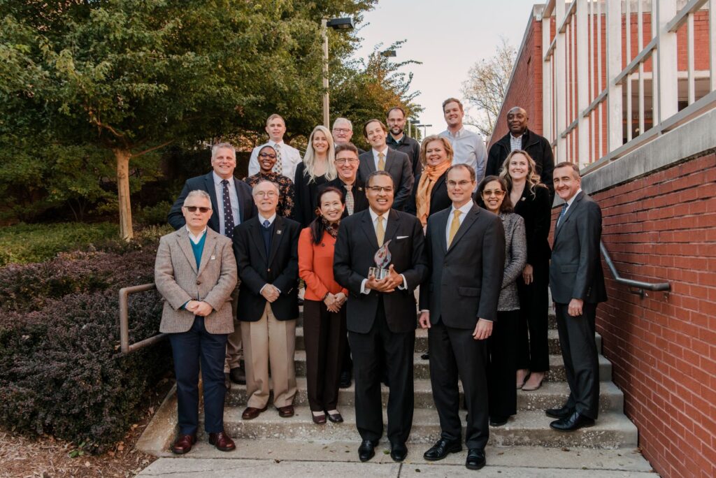 A diverse group of 19 adults in professional attire pose with a trophy.