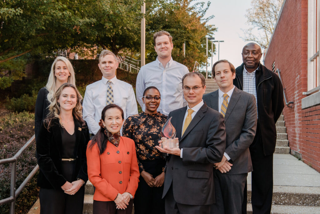 Nine people in professional clothing pose outdoors with a glass trophy shaped like a flame.