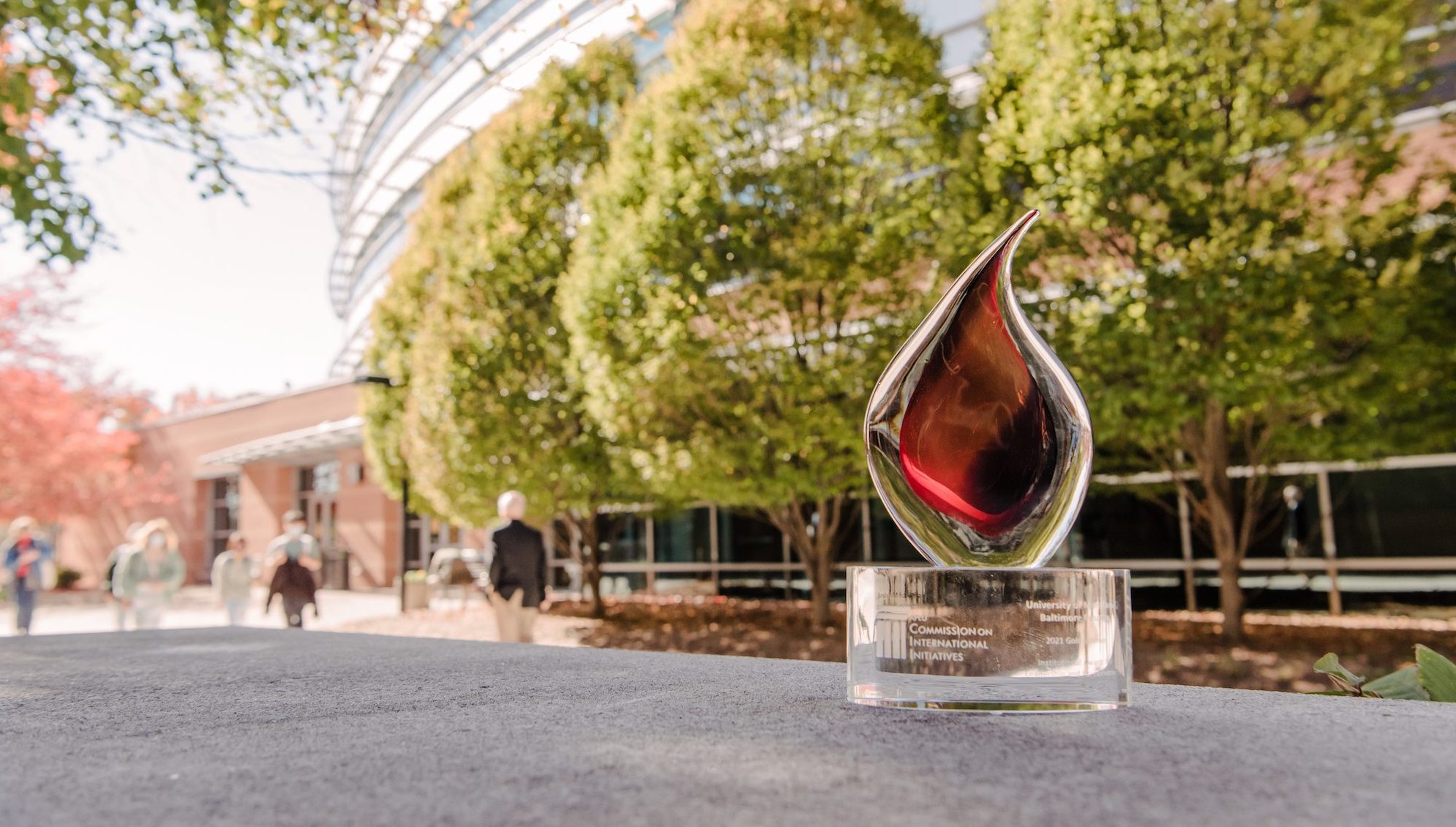 Glass trophy shaped like a flame rests on a concrete surface in front of trees and buildings
