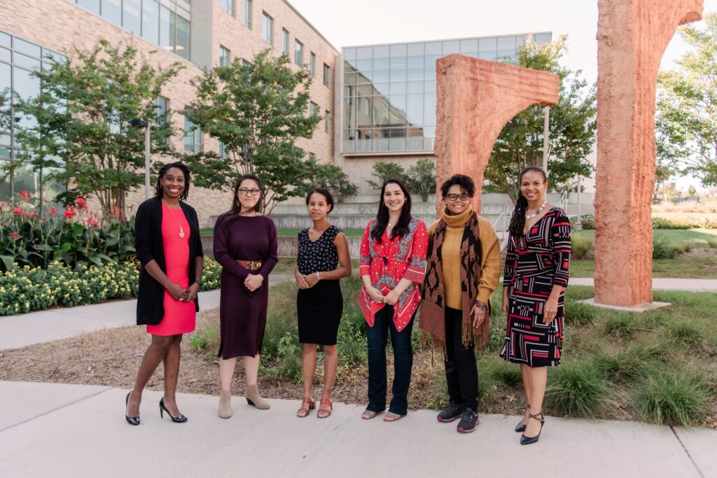 Five women wearing dress clothes stand in a group smiling at the camera. There are trees, large orange stone arches, and a a brick building with a glass wall building behind them.