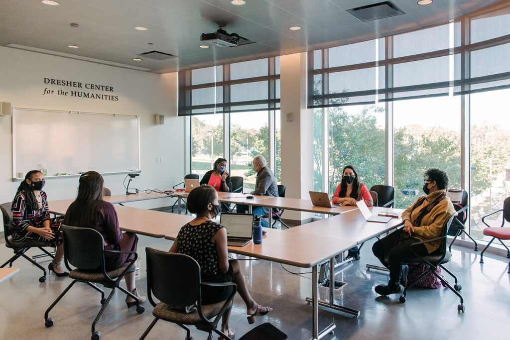 A group of seven people sit around a group of tables in a room with glass windows and a white board facing them.