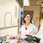 Woman with auburn hair stands with a man with dark bears in a lab. She wears a tie dyed lab coat and he wears a white lab coat.