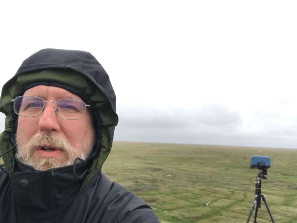 Man in hooded winter coat with green tundra, gray sky, and tripod in background.