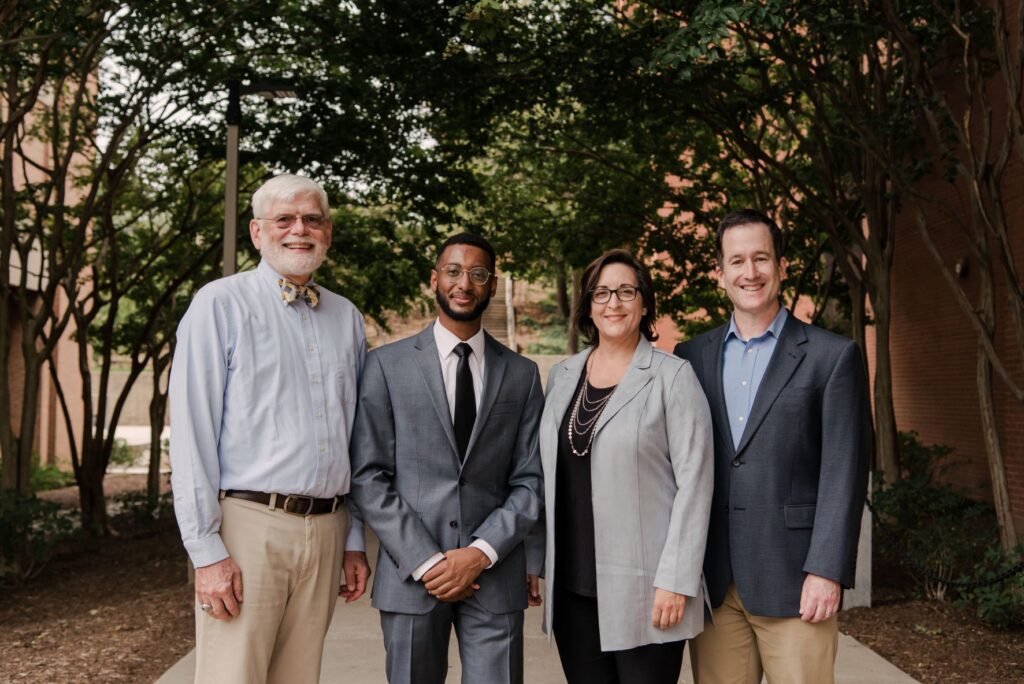 Four people wearing dress clothes stand next to each other on a path with trees and buildings in the background. They are all smiling. 