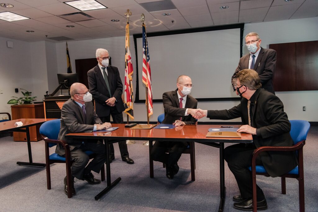 five people at a boardroom table, two shaking hands before signing documents before them.