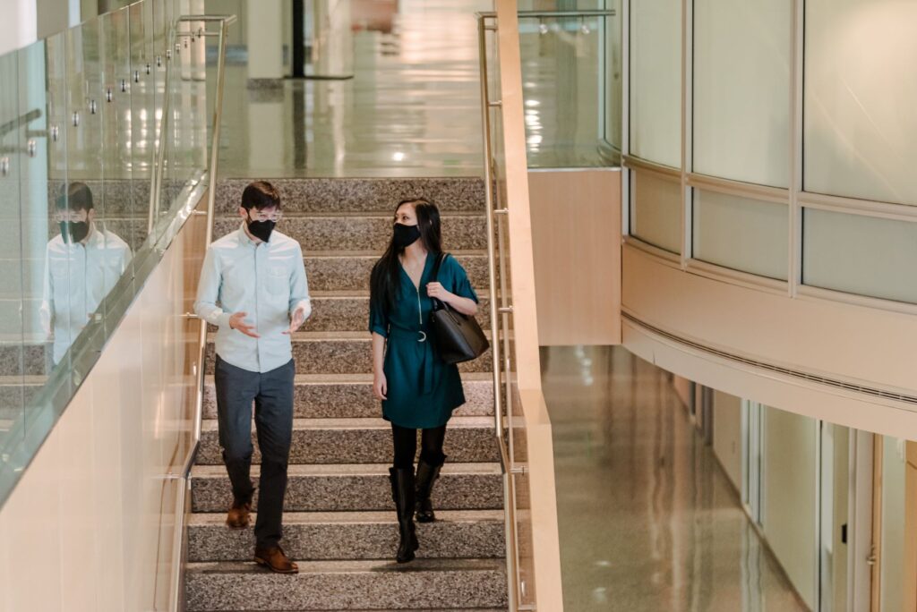 A woman in a blue dress and boots and man in button-up shirt talk, wearing face masks, as they walk down stairs inside a building.