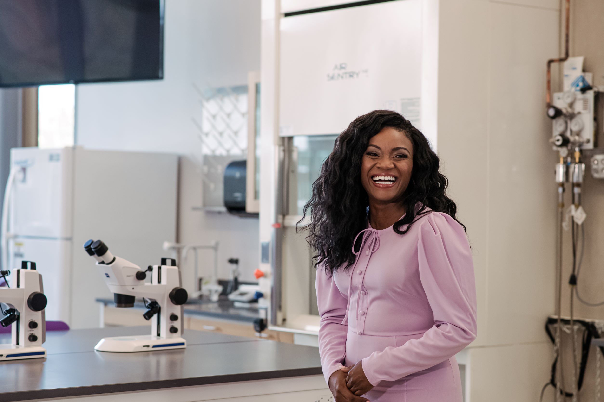 Black woman with long, curly hair smiles while standing next to microscopes in a lab.