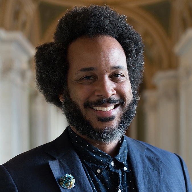 Headshot of smiling Black man with a beard, wearing a navy blazer with a flower pin.