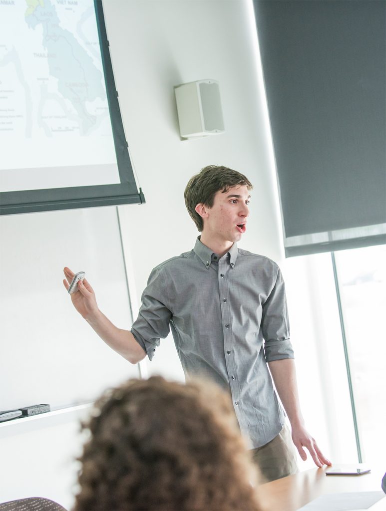 A young man stands in front of a projector screen displaying a map. He is giving a presentation and has his hand up, holding a device to flip through his presentation slides.