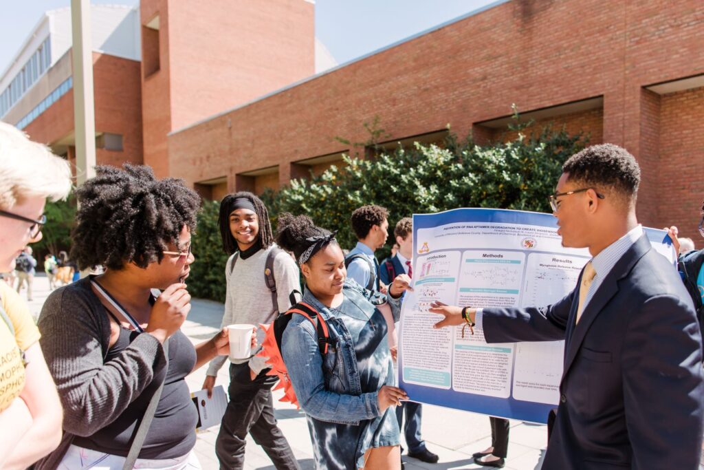 A student in a suit and glasses points to a presentation poster, held by another student, outside of an academic building.