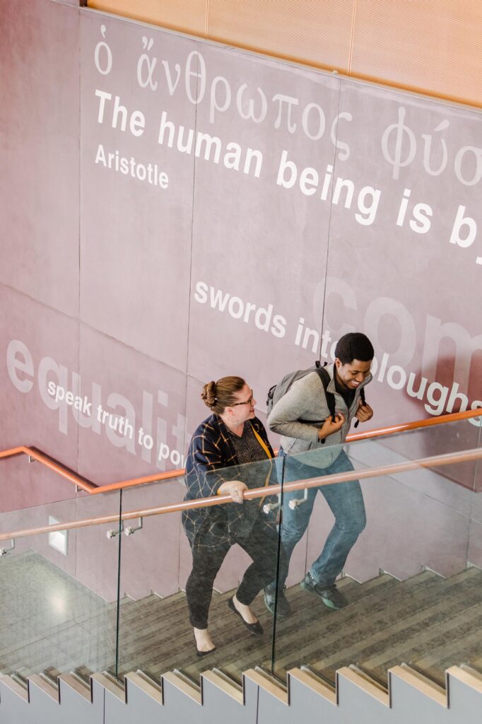 A woman and young man walk up a flight of stairs. Next to them is a large wall with graphic text on it, displaying a quote by Aristotle. Only part of the quote is visible, reading "The human being is..."