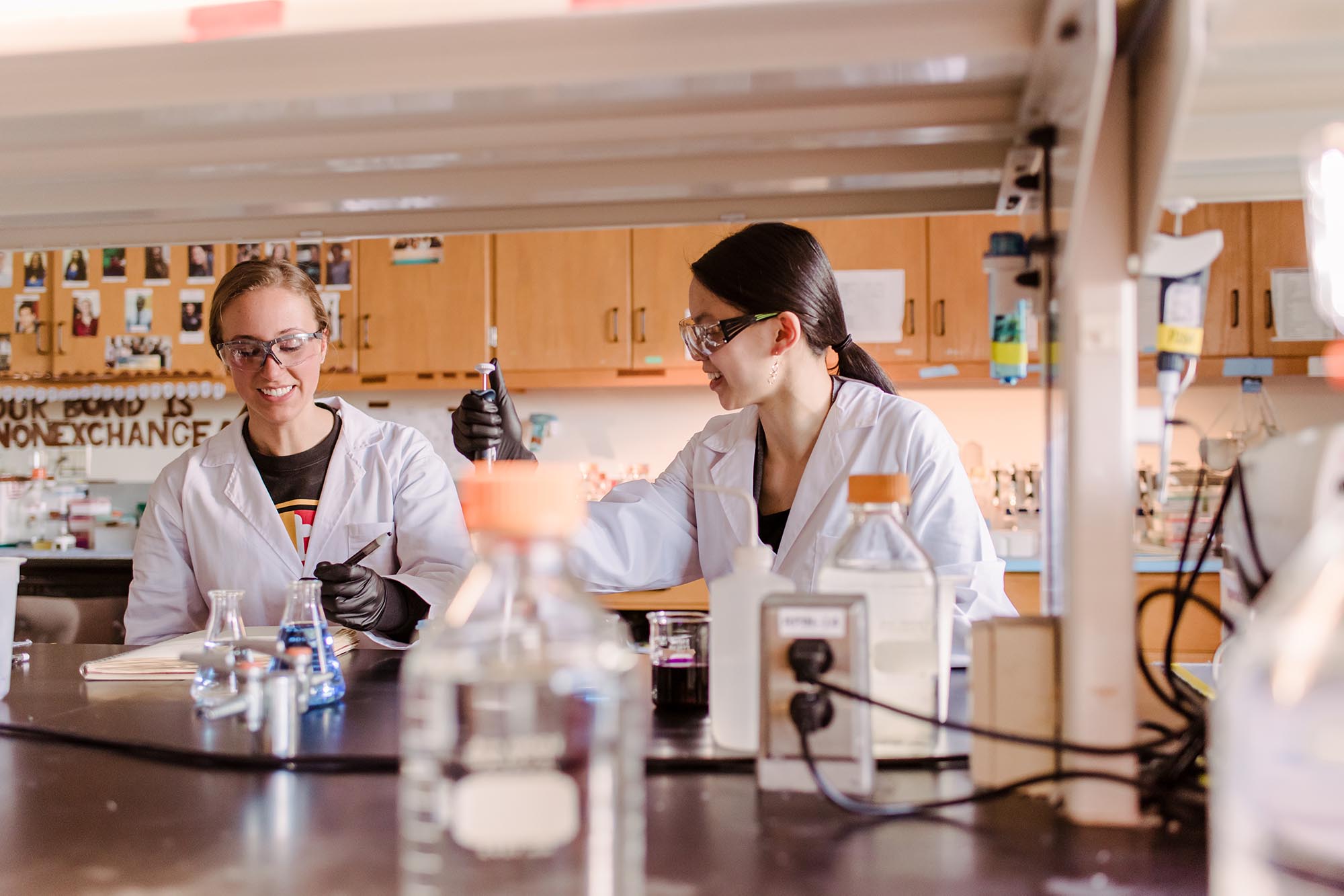 Two female students wearing white labcoats and protective eyewear conduct laboratory research at umbc.