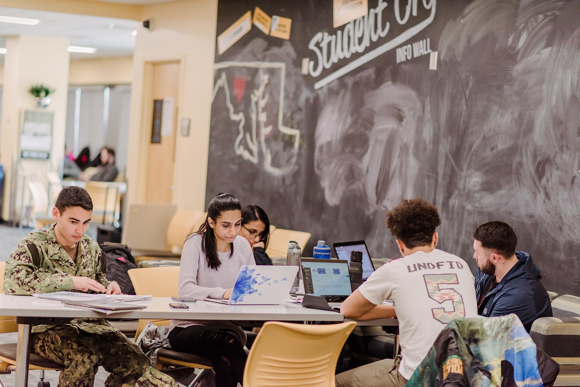 A diverse group of five young women and men study together. Each student has a laptop, and behind them is a large chalkboard.