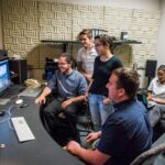 Four students gather around monitors, with a professor and student sitting in the background.