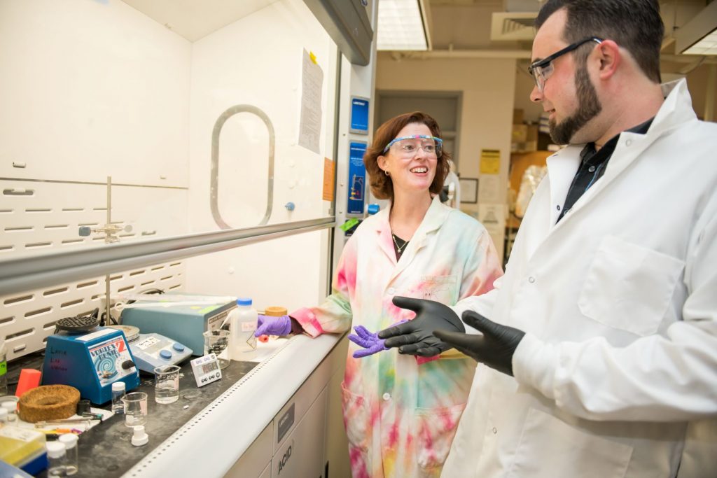 Woman with auburn hair stands with a man with dark bears in a lab. She wears a tie dyed lab coat and he wears a white lab coat.