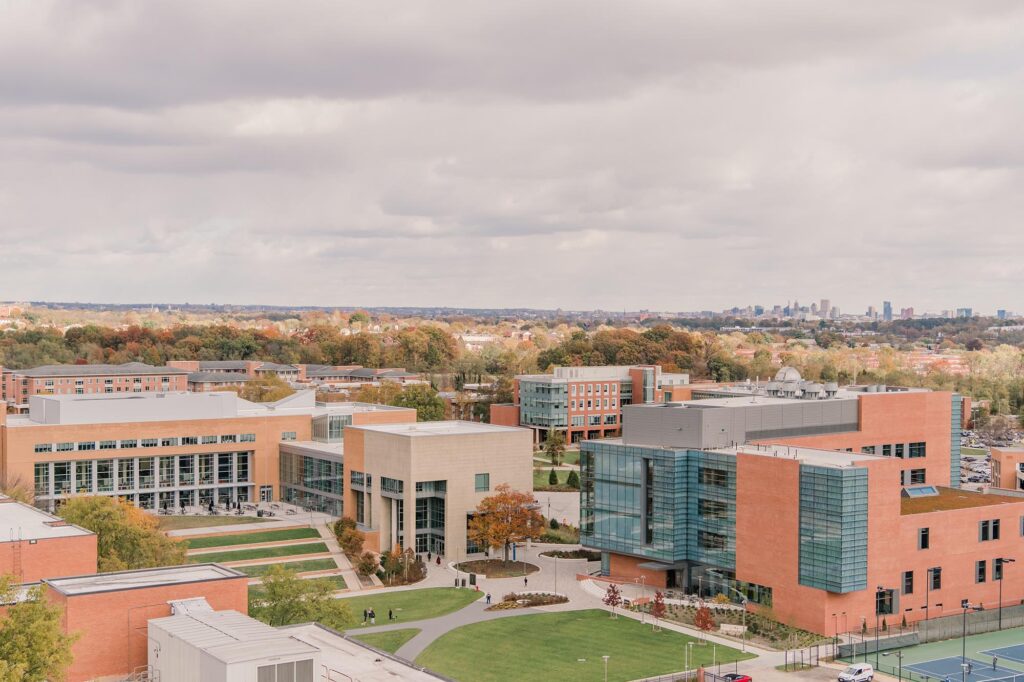 Aerial view of the UMBC campus on an overcast day, showcasing modern red brick buildings with glass facades, manicured green lawns, and walking paths. In the distance, a panorama of autumn-colored trees and the Baltimore city skyline.