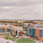 Aerial view of the UMBC campus on an overcast day, showcasing modern red brick buildings with glass facades, manicured green lawns, and walking paths. In the distance, a panorama of autumn-colored trees and the Baltimore city skyline.