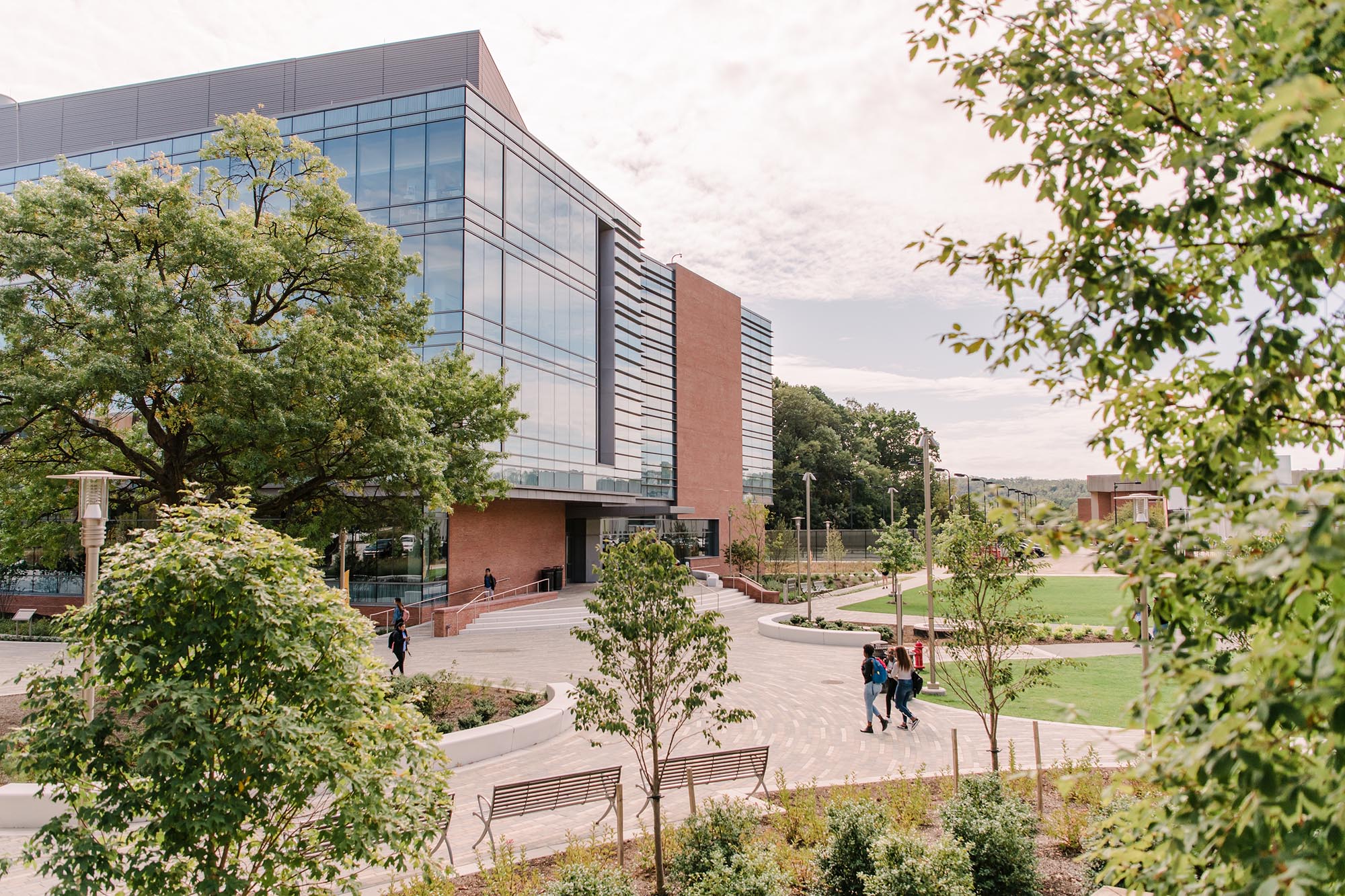 UMBC's campus becomes green in the Spring season. Students enjoy the trees and fresh air while walking past a unique brick building with large glass windows.