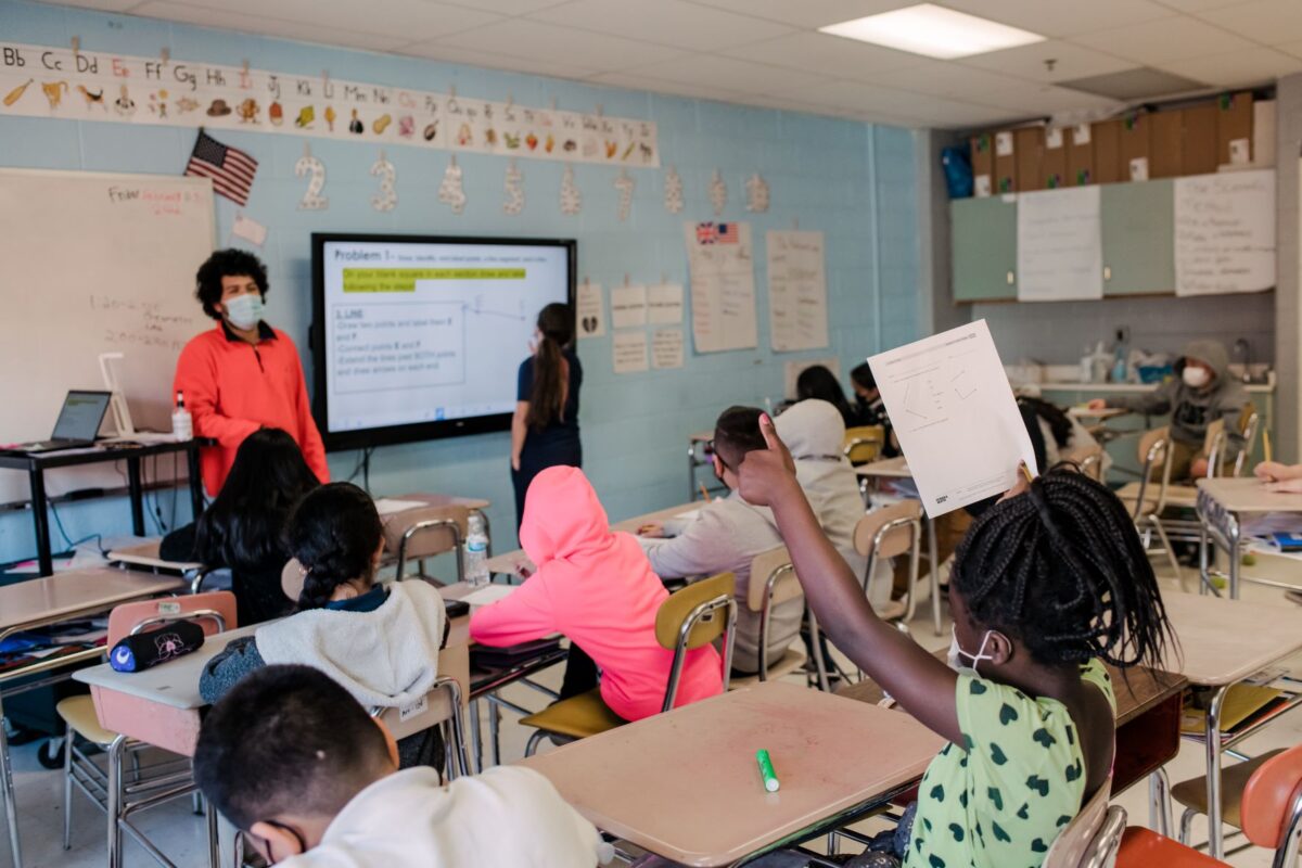 Student raises her hand while seated in class, surrounded by other students, with teacher at the front of the room.