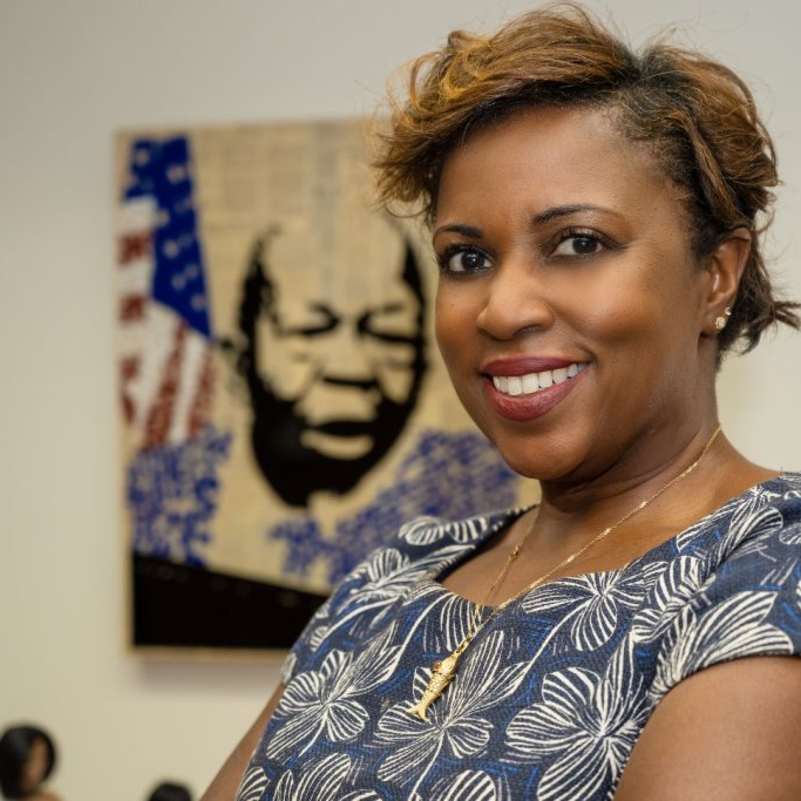 A woman wearing a blue and white floral shirt and gold necklace with a portrait of a man with the American flag behind her.