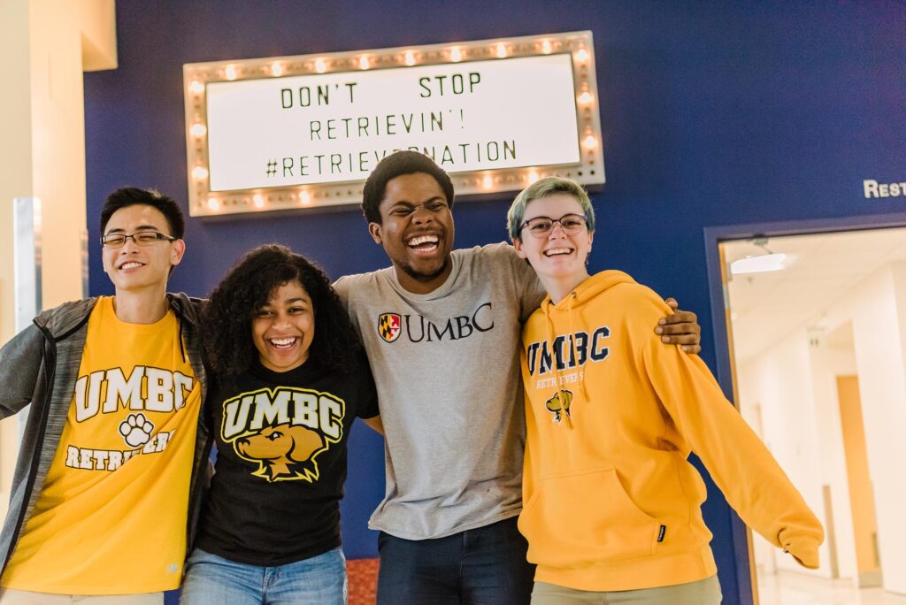 Four joyful friends, each wearing a different UMBC shirt, stands in front of a brightly lit event sign that reads, "Don't Stop Retrievin' hashtag Retriever Nation."