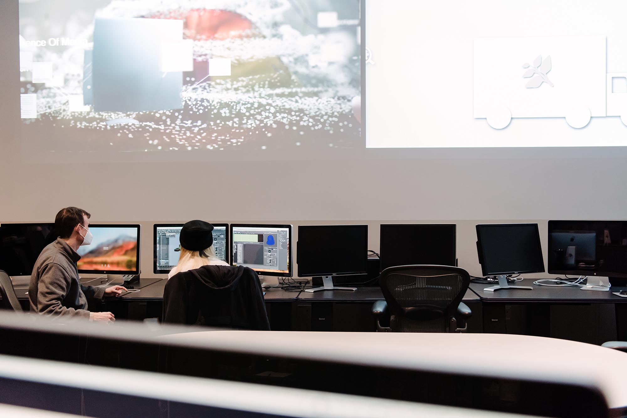 Two people working in a computer lab together, a projector is showing imagery above them.