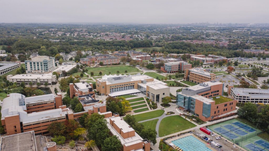 A view of the UMBC campus from above, taken from a drone. The campus buildings and fields are visible, as well as the skyline of nearby towns in the distance.