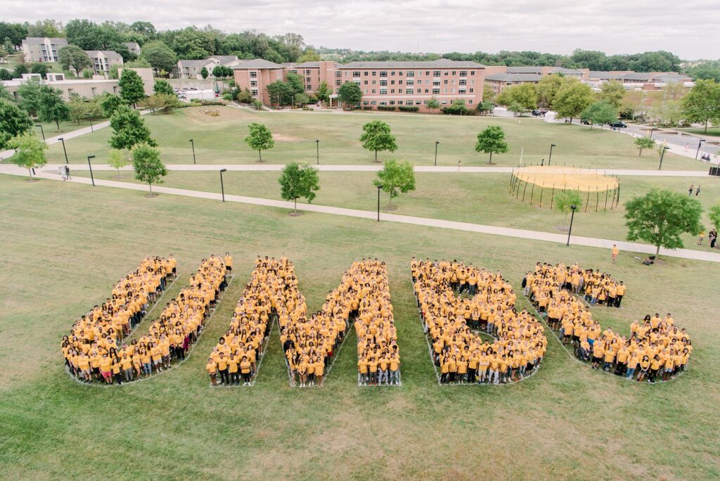 A large body of students standing in groups that spell out UMBC when seen from bird's eye view. They are wearing gold on top, and black bottoms to represent school spirit.