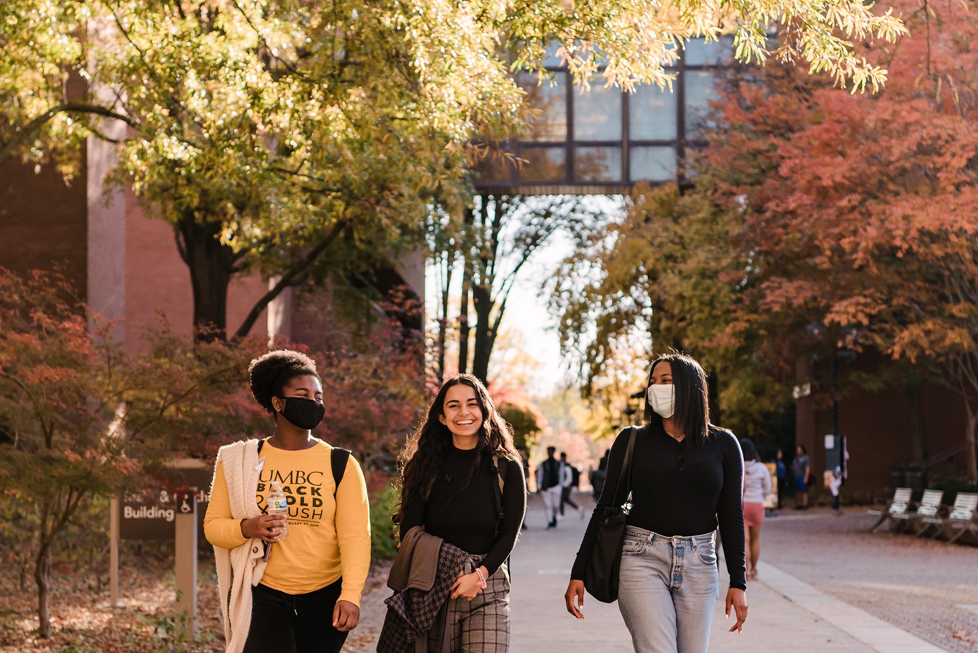 Three young women walk along UMBC's academic row, having a joyous conversation. Two are wearing masks, one has a shirt that reads "UMBC Black and Gold Rush."