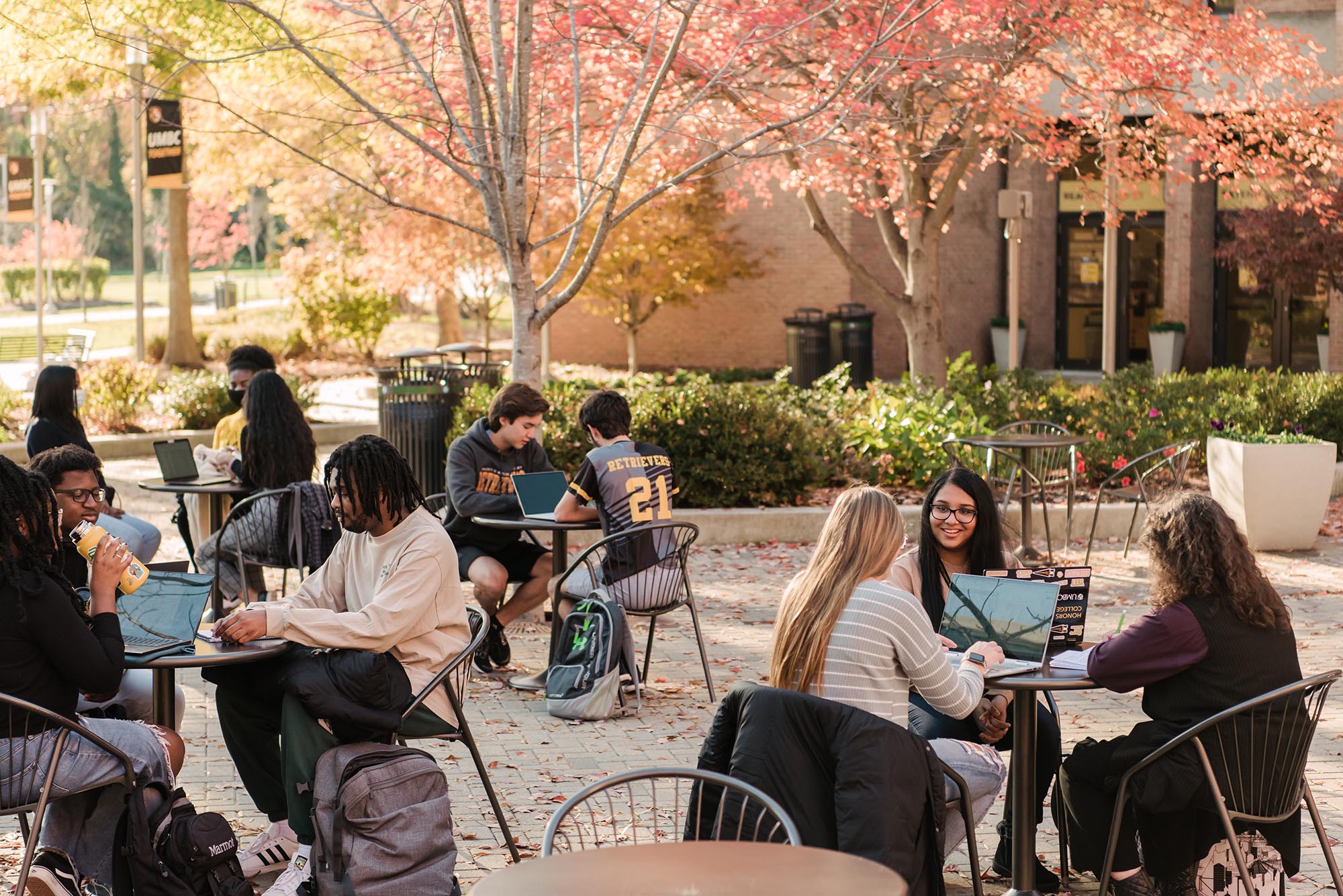 A diverse group of young students sit outside at different tables around the university center. Some students are working on their laptops, some are chatting and drinking water. Buildings and trees surround them in the background.