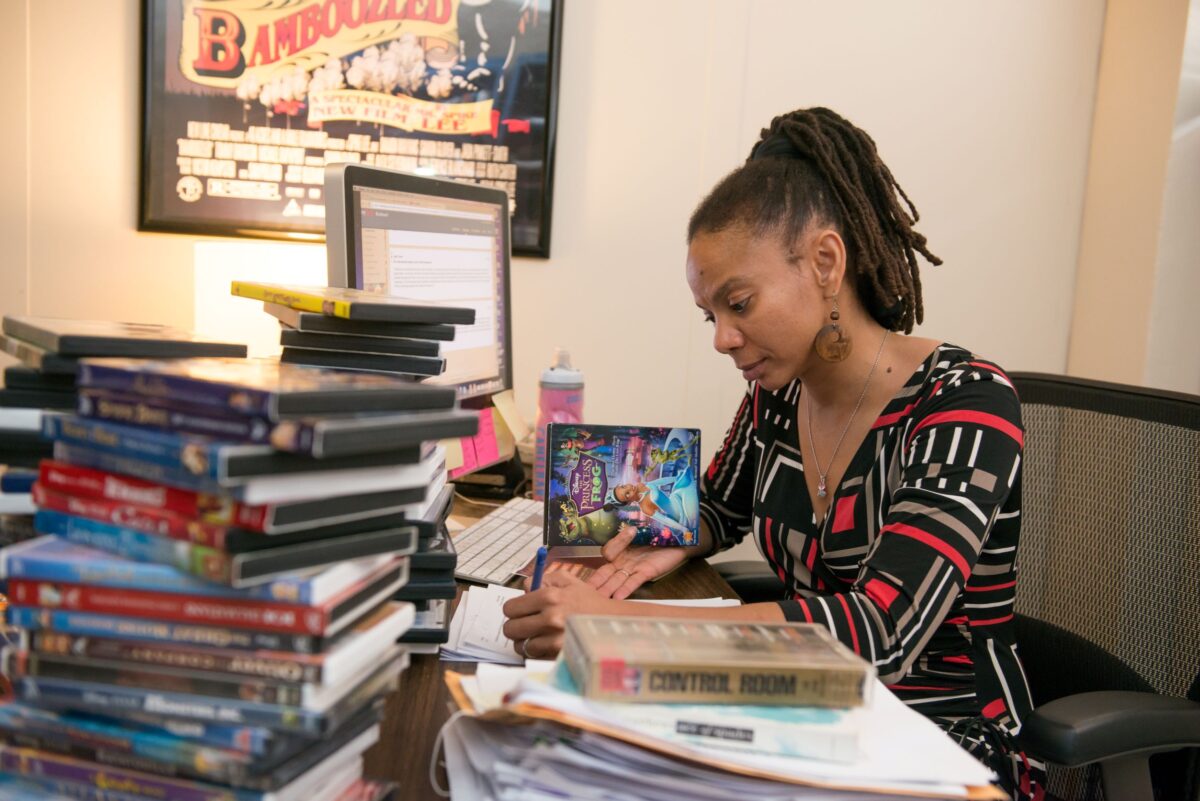 A woman sitting in an office at a desk featuring piles of DVDs.
