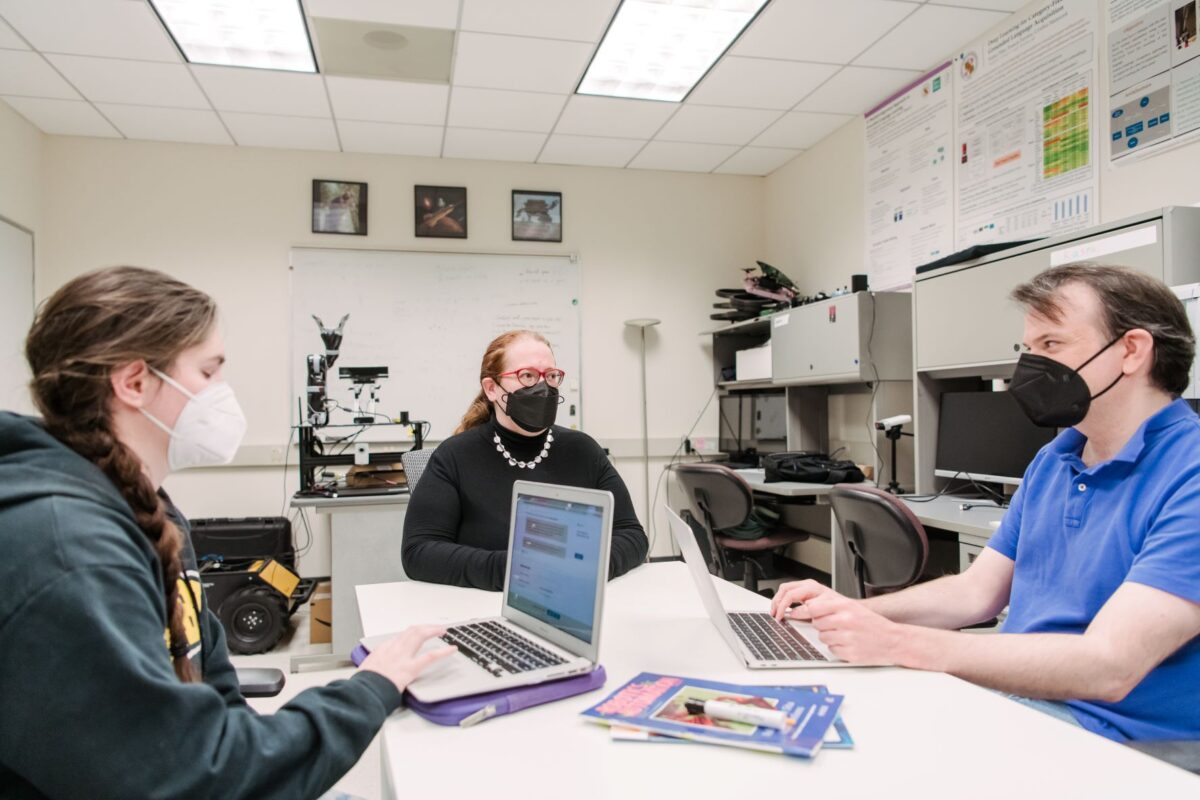 Cynthia sits at the end of a table with two other people looking at their laptops. Everyone has a mask on.