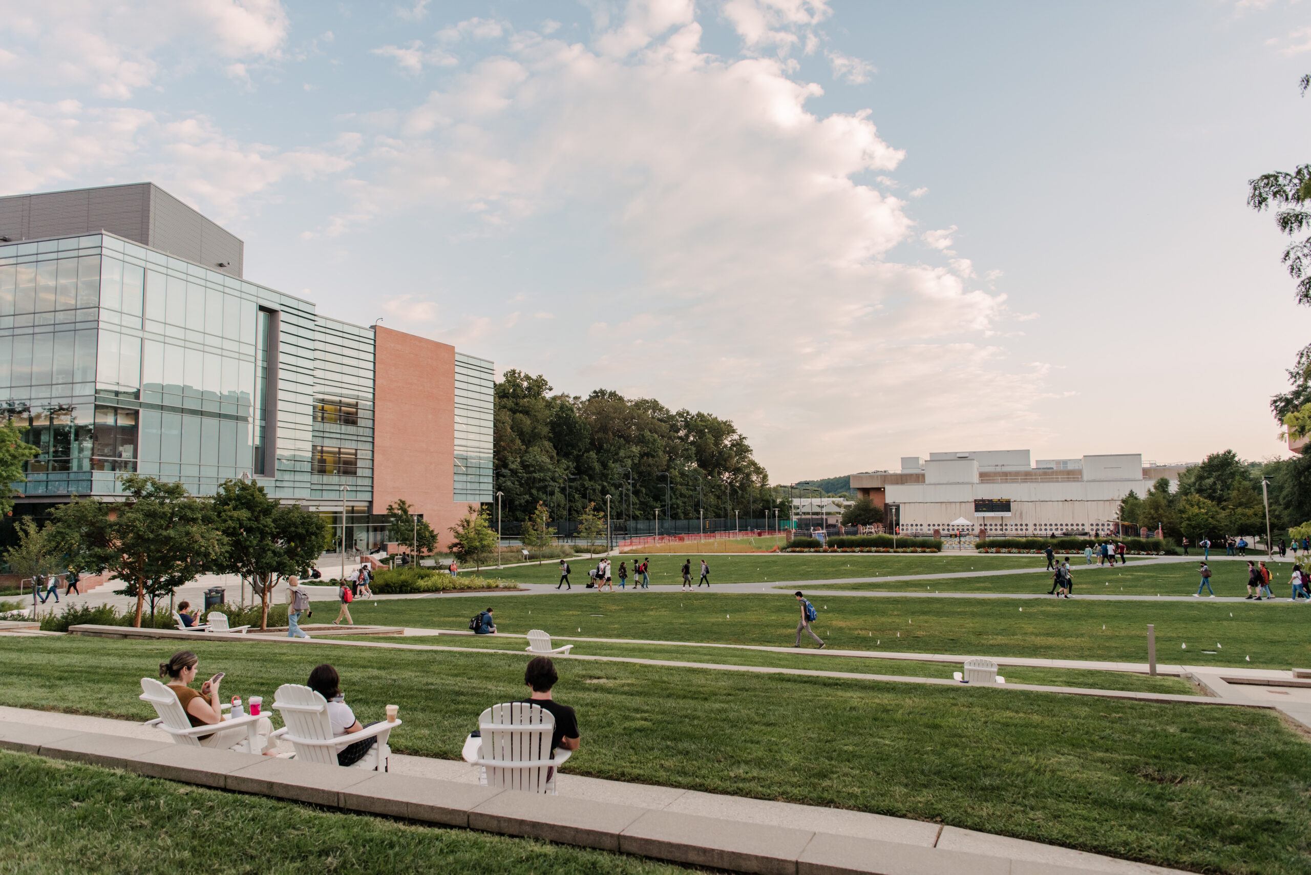 Three friends sit together on white lawn chairs and overlook the campus quad, which is lush and grassy. Many students walk around in the distance, and there are a few buildings and trees around them.