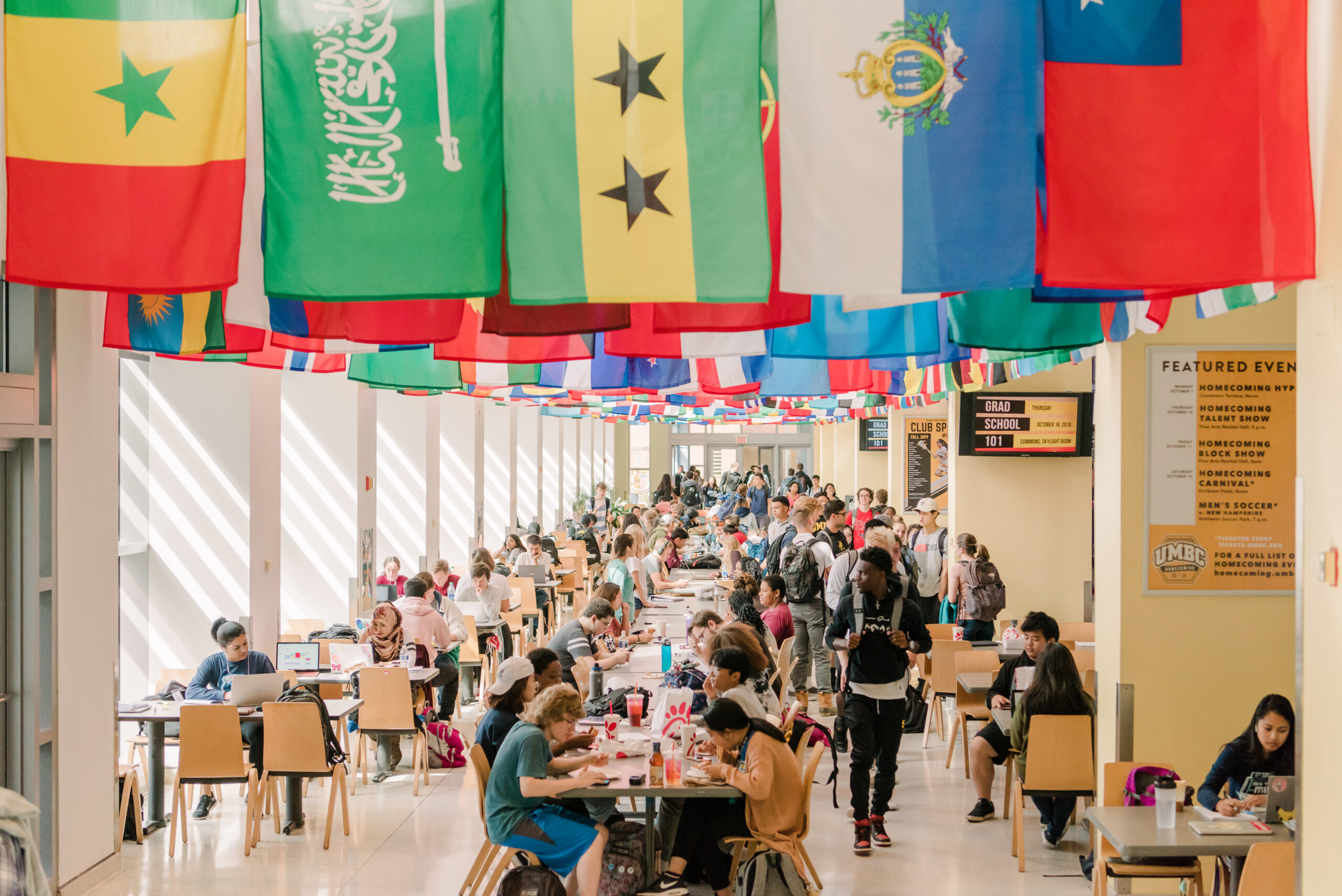 UMBC's commons is bustling with a diverse community of students. Some are on their way to class, some are eating, and some are working on assignments. Above them hangs a large collection of all the country flags. Posters on the walls display upcoming featured events, club sports, and information about grad school.