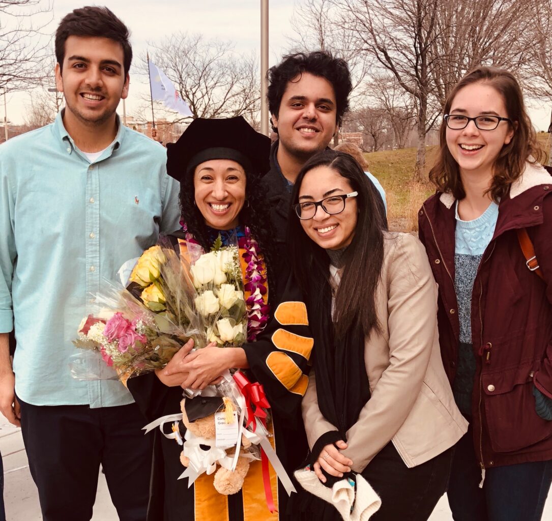 Four undergraduates with Mariann Gabrawy, who is dressed in commencement regalia and holding several flower bouquets. 
