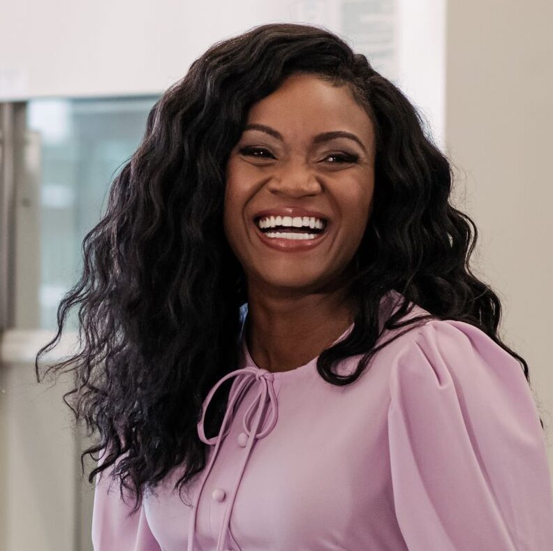 Black woman with long, curly hair smiles while standing next to microscopes in a lab.