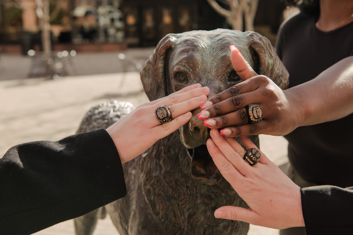 UMBC's mascot is a Chesapeake Bay Retriever named True Grit, and a well-known statue of him sits in front of the campus gym. Three young women hold their hands over his nose for good luck. Each hand is wearing an alumni ring.