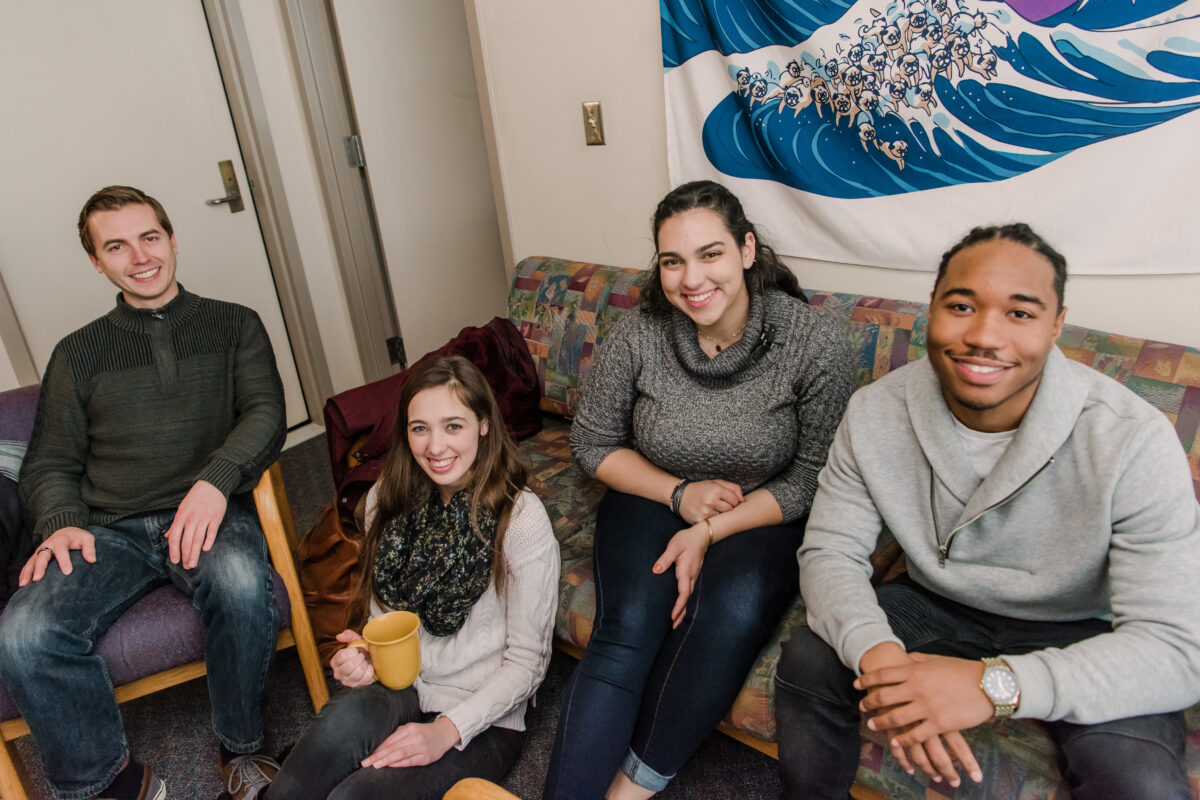 A group of four diverse young women and men sit together in a dorm room.
