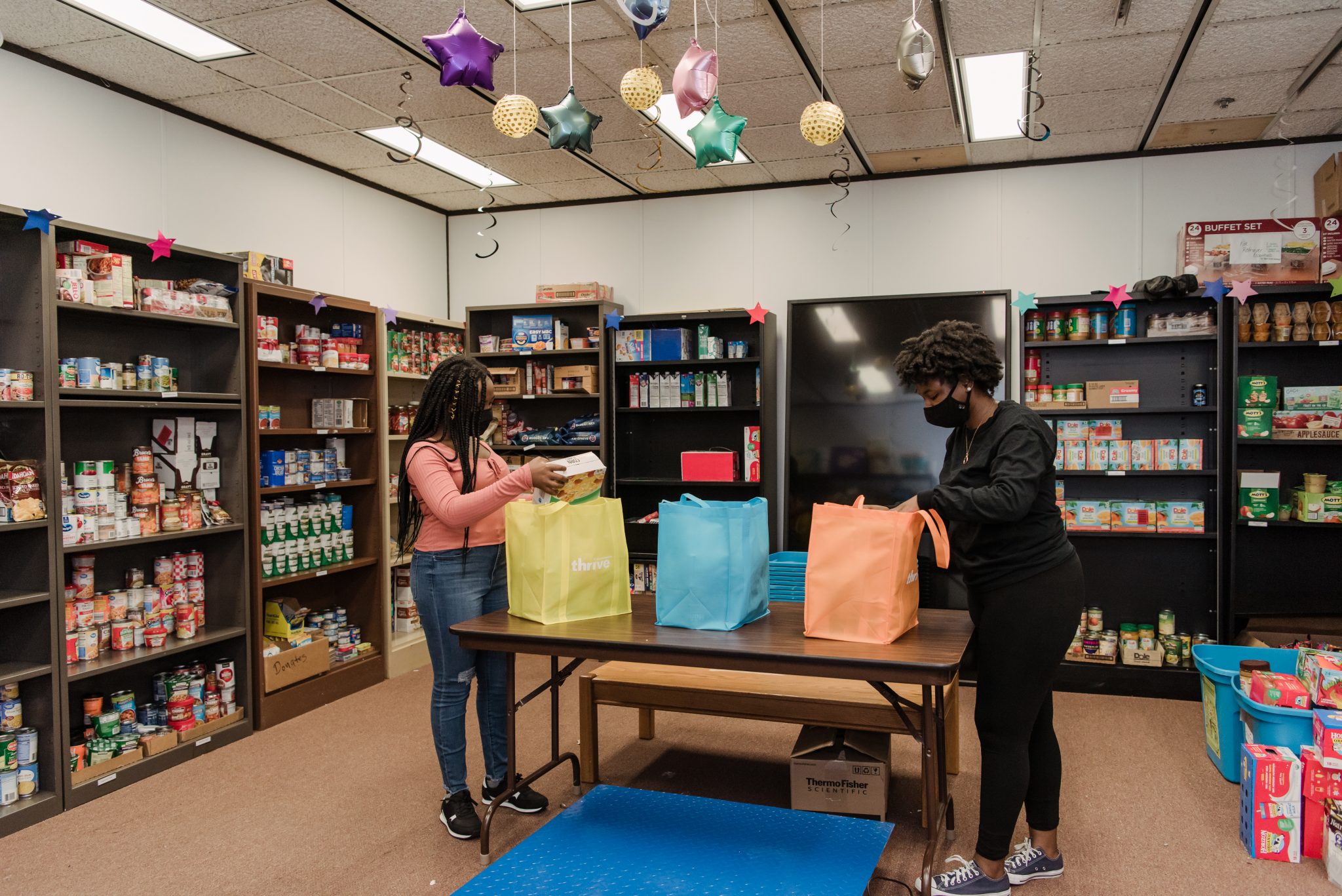 Two students are packing three bags full of donated food. They are standing in a pantry with many shelves full of canned foods and goods.