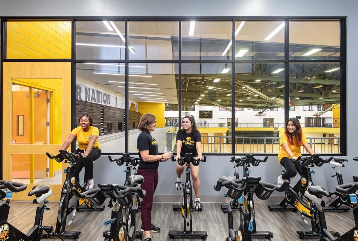A group of four women using exercise bikes, with a window behind them that overlooks the RAC's running track.