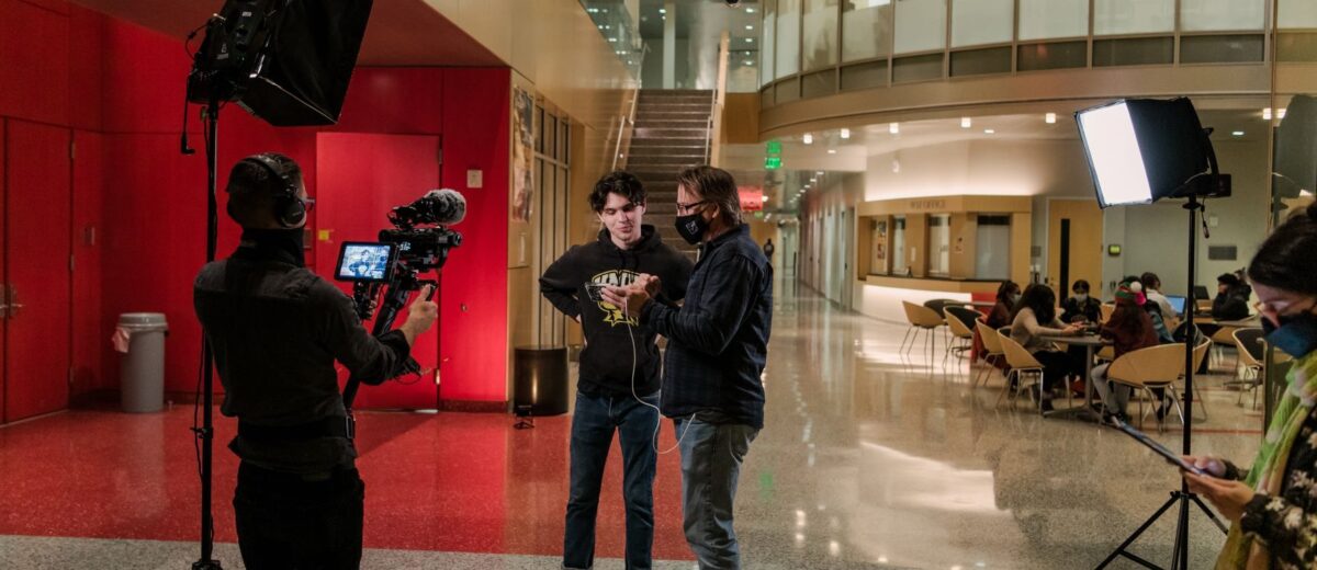 A student stands looking at a phone screen with a man while a camera operator looks on.