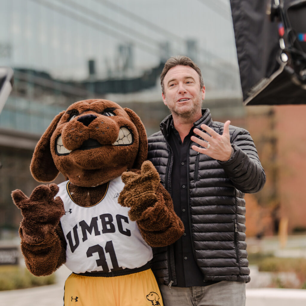 The College Tour host Alex Boylan poses with UMBC Mascot True Grit. 