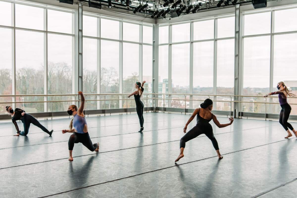 Five elegant dancers wearing leotards are posing in a dance studio with glass windows looking over the UMBC campus.