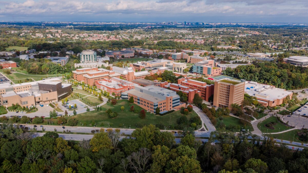 Aerial drone shot of UMBC campus full of bright brick buildings. The Baltimore skyline can be seen in the distant horizon.