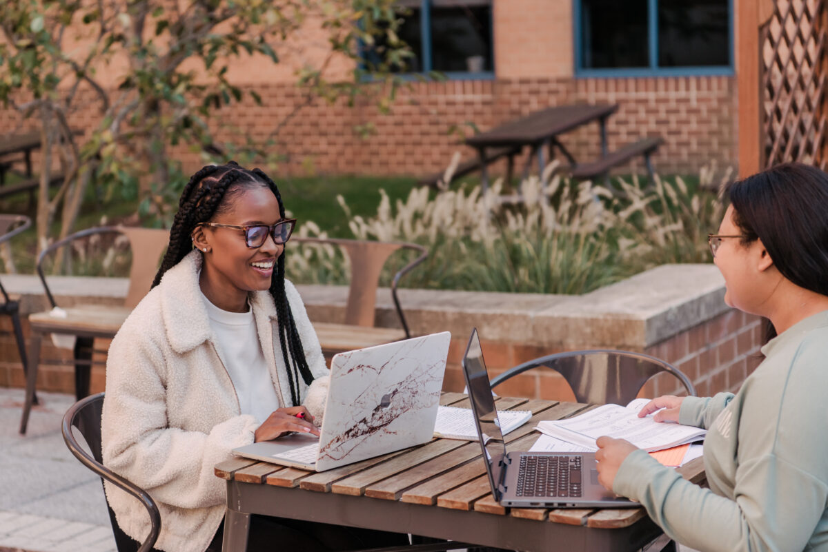Two young ladies sitting outside a residential area, smile at each other and use their laptops. Behind them is a brick building with trees and plants.