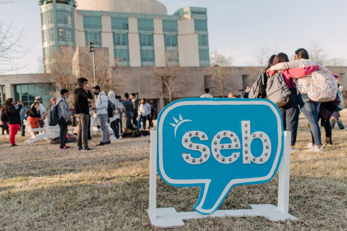 A crowd of people stand outside of the Albin O. Kuhn library, with a blue SEB sign in front of them.