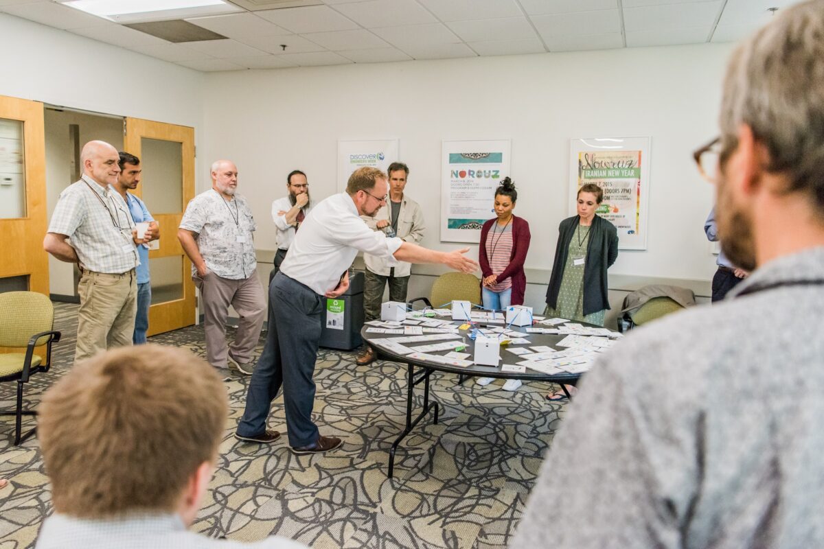 adults stand in a circle; one in the center is pointing to notecards laid out on a table